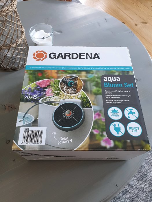 Gardena Aqua Bloom Set för bevattning ligger oöppnat på ett bord med inredningsdetaljer i bakgrunden.