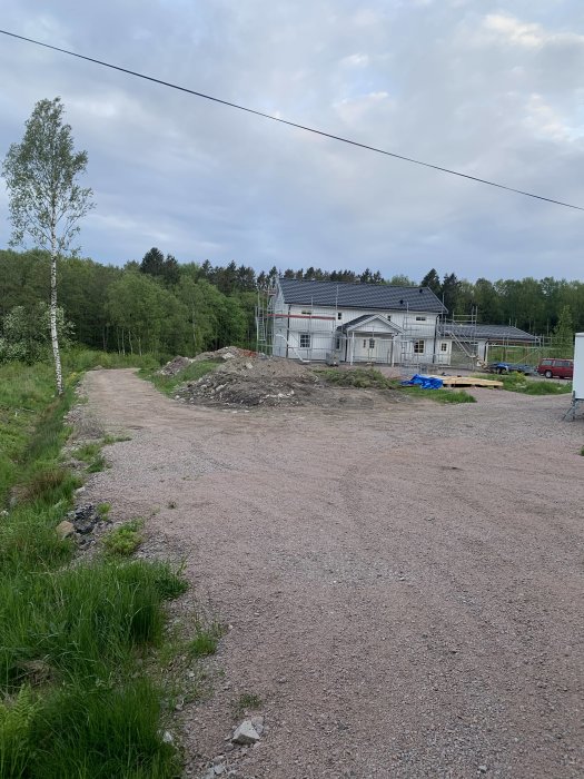 Byggarbetsplats med halvfärdigt hus och byggställningar, omgivet av natur och grusväg.