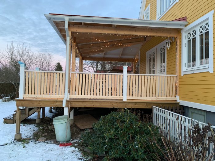 Nybyggt trästaket med planhyvlade spjälor på en veranda i anslutning till ett gult hus, omgivet av vintersnö.