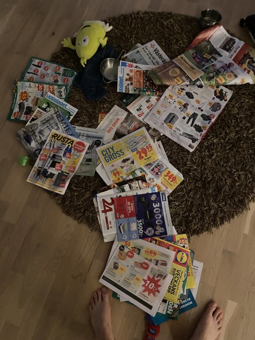 Reklamblad och kataloger utspridda på ett golv med en leksaksfigur och ett barns fötter synliga.