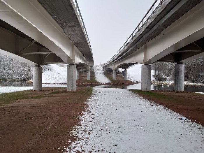 Två parallella motorvägsbroar av betong över en flod med lätt snötäcke på marken.