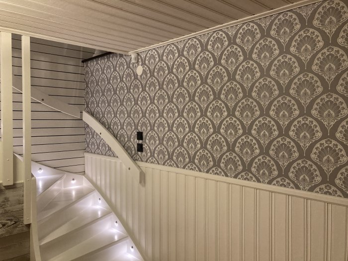 Renoverad trappa med LED-belysning, vit panel och mönstrad tapetvägg.