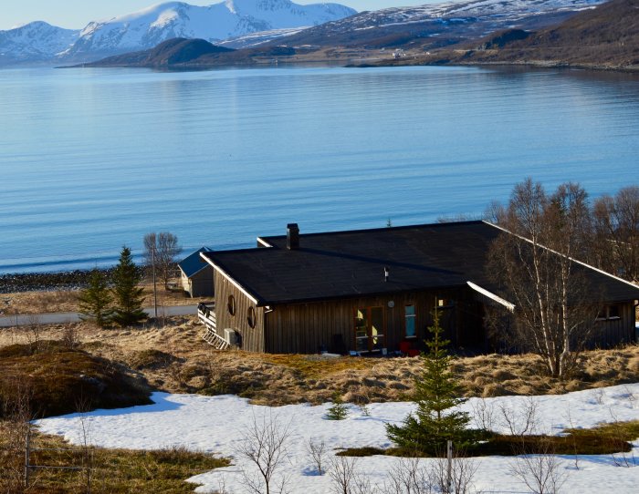 Stuga med svart papptak i norskt landskap med hav och snöklädda berg i bakgrunden.