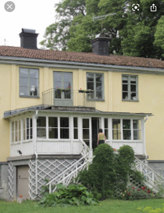 Gul villa med vit glasveranda, trästaket och trappa omgiven av grönska.