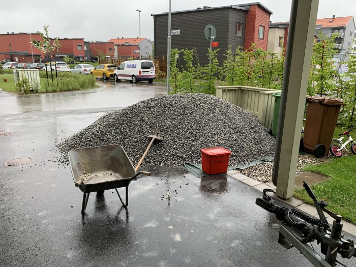 Stor hög med makadam på uppfart, skottkärra och regnväder, bostadshus i bakgrunden.