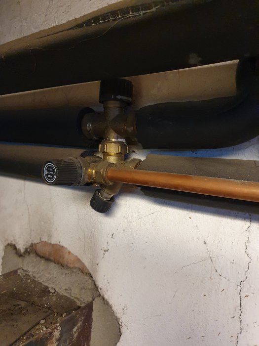 Värmesystem med isolerat svart varmvattenrör och förgreningsventil på vit vägg.