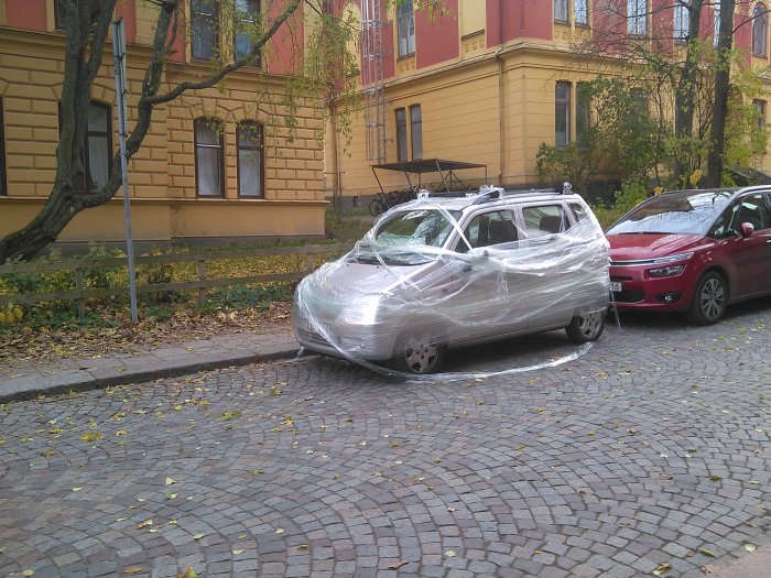 Bil insvept i plastfolie parkerad mellan andra bilar på en gata.