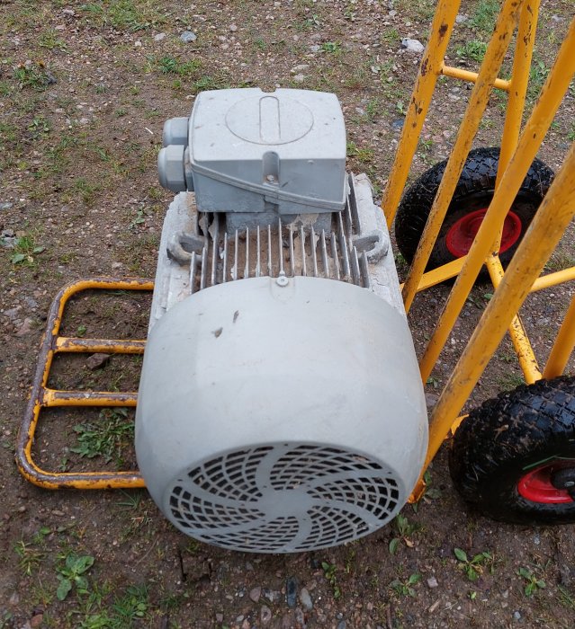 En 18.5kw grå industriell motor ligger på marken bredvid en gul hjulburen handkärra.