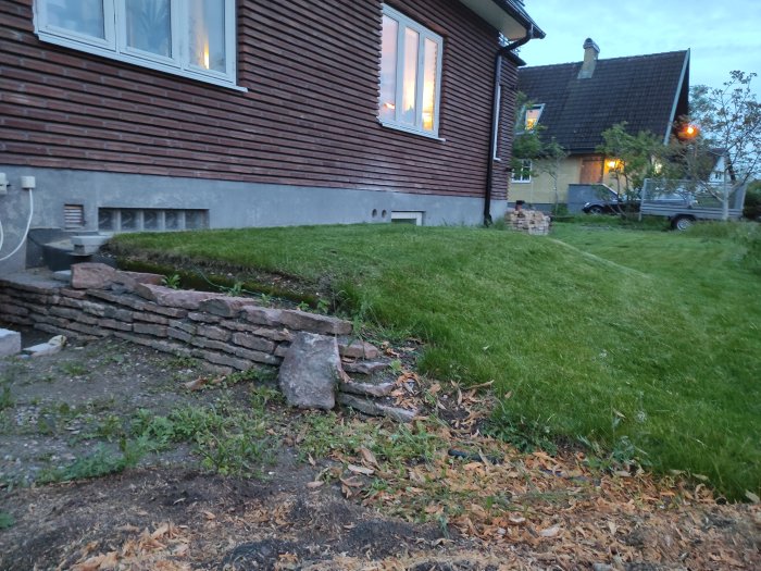 Nybyggd gräsbevuxen gjuten platta vid husgrund med synlig armering och stenmur i förgrunden.