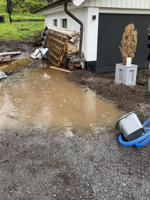 Översvämmad trädgård nära garage med synliga fördämningar och en pump.