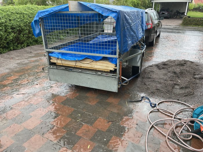 Släpvagn med presenning i regn, våt uppfart, byggmaterial, bil och högar med grus.