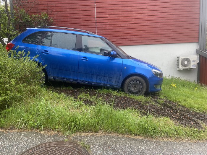 Blå bil parkerad på sluttande gräsmatta med täckbark för att förhindra lera.