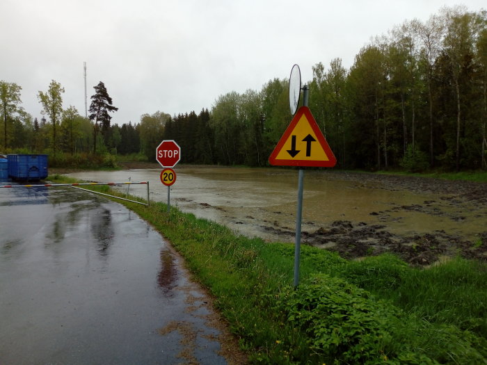 En översvämmad byggarbetsplats med stoppskylt och varning för vägarbete, omgivet av träd.