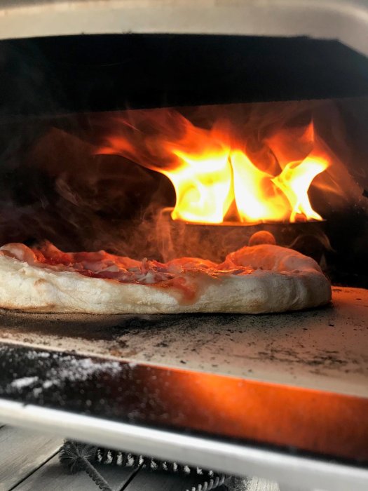 Hemlagad pizza inne i en vedeldad Ooni pizzaugn med synliga lågor i bakgrunden.