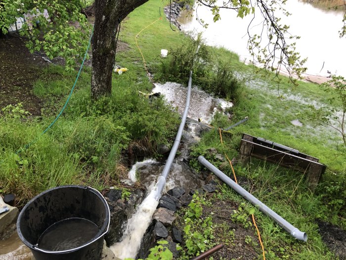 Översvämmad trädgård med rör och hink används för att leda bort vatten mot havet.