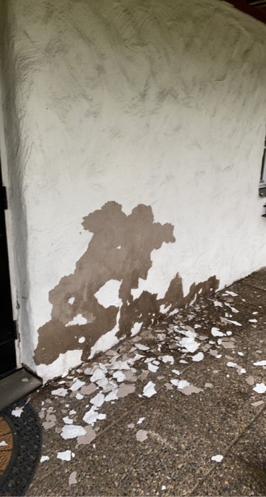 Vit fasad med flagnad puts som avslöjar underliggande mörkbrunt material, samt putsbitar som ligger på marken.