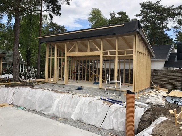 Byggplats med trästomme för hus, halvt komplett tak och material utspridda på marken.