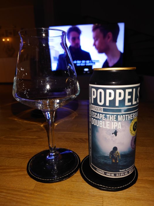 Ölburk från Poppels Bryggeri och tomt ölglas på bord framför TV som visar en film.