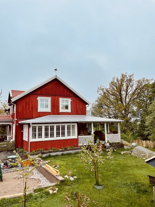 Rött hus med vit veranda runt hörnet och pågående trädgårdsarbete i förgrunden.