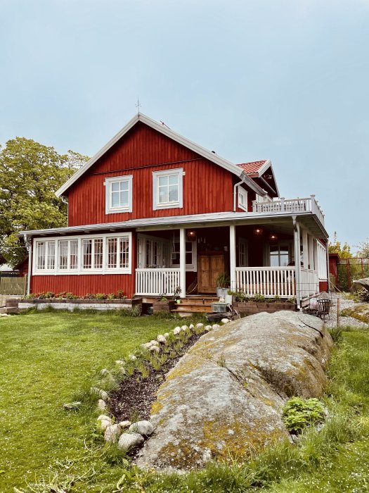 Rödmålat trähus med veranda som går runt hörnet, beläget i en lummig trädgård med stenformation framför.