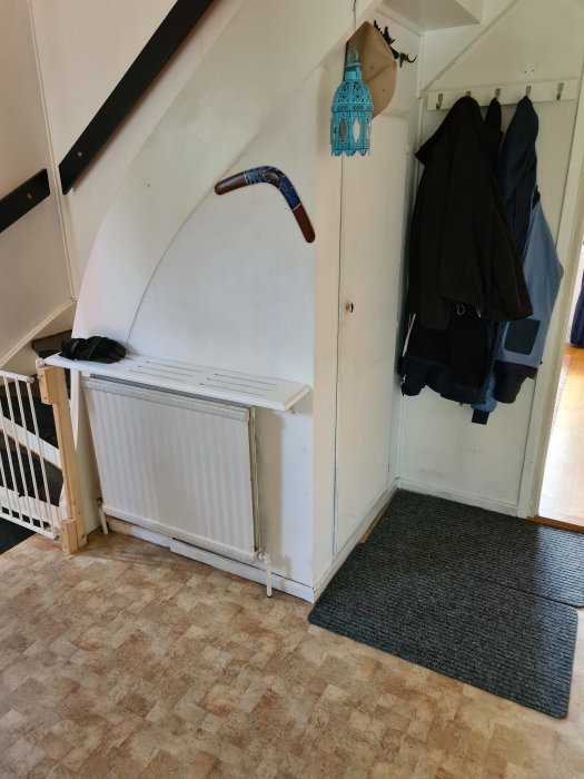 Hall med vit vägg under trappa, vattenburen radiator och klädhängare samt ett litet skåp.