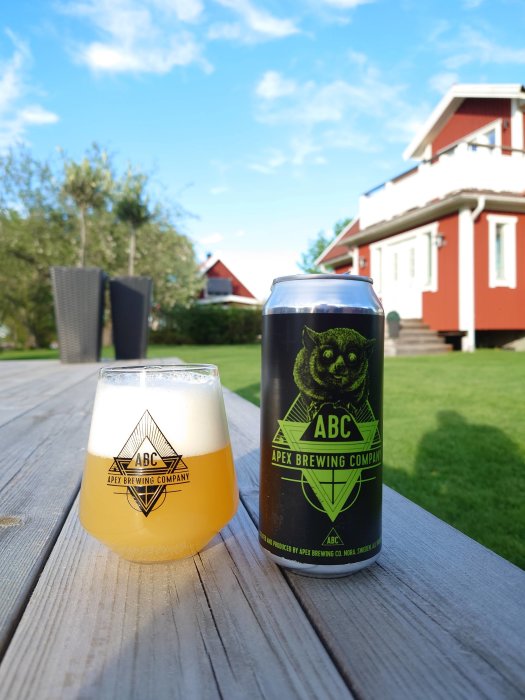 Ölglas med skummande dryck och ölburk från APEX Brewing Company på träbord utomhus med hus i bakgrunden.