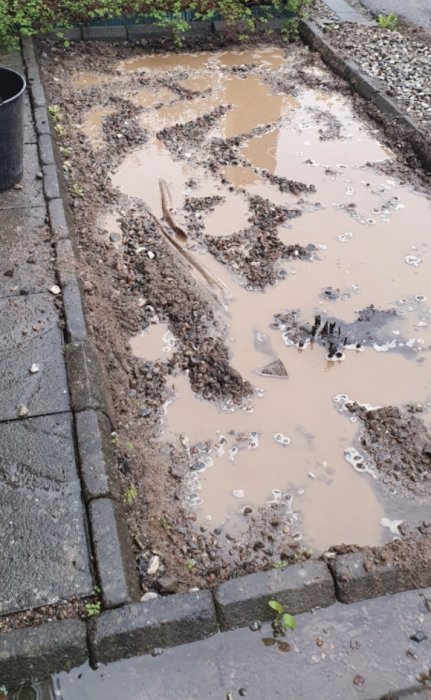 Grävd plats med borttaget grus och sand, vattensamlingar och smutsig markduk i ett trädgårdsområde.