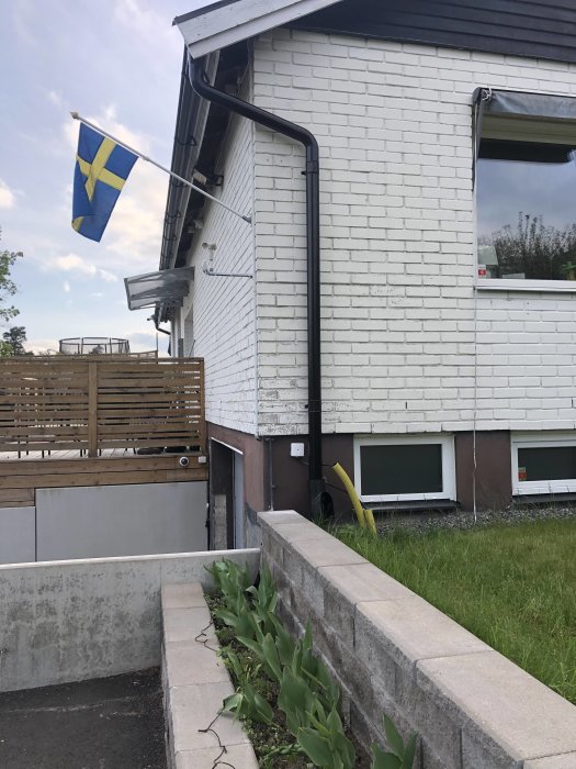 Flyttat stuprör vid hörn av vit tegelhus med nytt svart staket och svensk flagga, redo för målning och generatorbyte på bil.