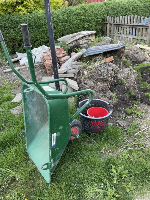 Grön skottkärra med ett rött däck framför en hög med jord och byggmaterial på en trädgård.