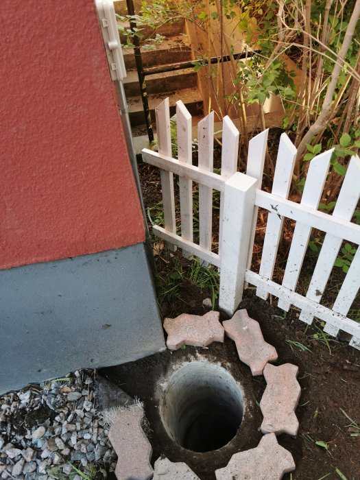 Betonglock täcker en dagvattenbrunn vid husrundan, omgivet av grus och ett vitt staket.