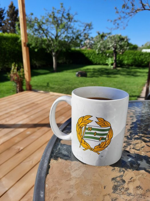 Kaffekopp med emblem på ett soligt trädäck med grönskande trädgård i bakgrunden.