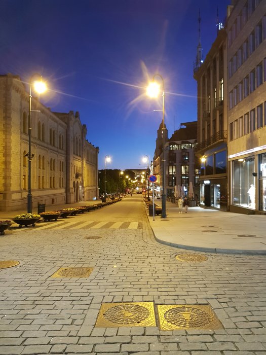 Kvällsbild av en öde Karl Johans gate i Oslo med gatubelysning och tomma trottoarer.