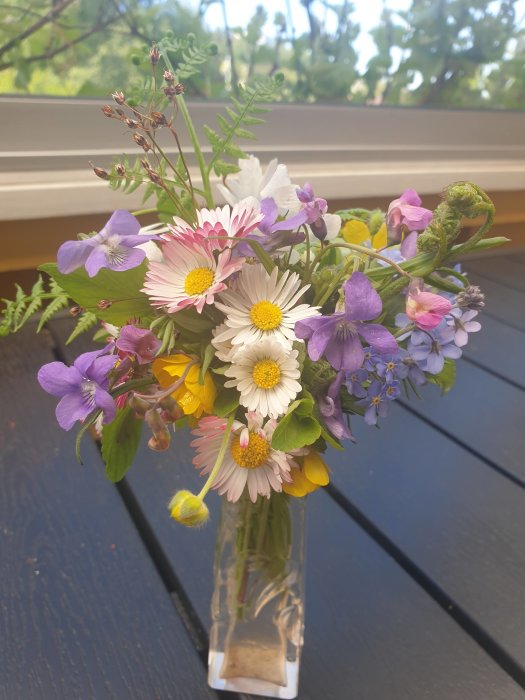 Bukett med blandade vårblommor i glasvas på svart träbord utomhus.