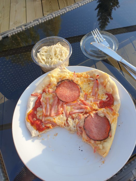 Hemlagad pizza med salami på vit tallrik bredvid en gaffel och coleslaw i plastburk på glasbord.