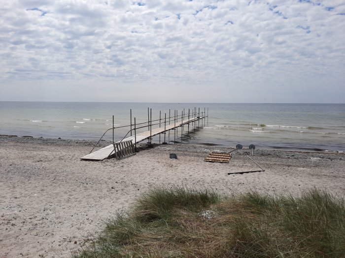 En nybyggd brygga som sträcker sig ut i havet från en sandig strand, byggmaterial syns på land.