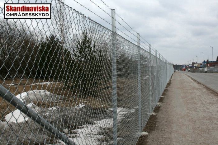 Kedjestängsel längs trottoar med företagets logotyp, i ett industriområde med snöfläckar.