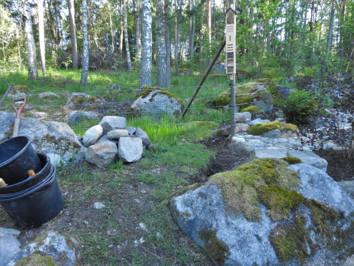 En trädgård under arbete med spade, hinkar, stenhög och ett nyligen uppsatt bihotell på en stolpe.