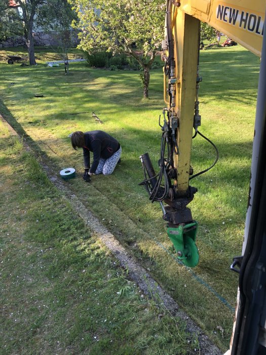 En person installerar begränsningskabel för robotgräsklippare i en trädgård medan en grävmaskin är i förgrunden.