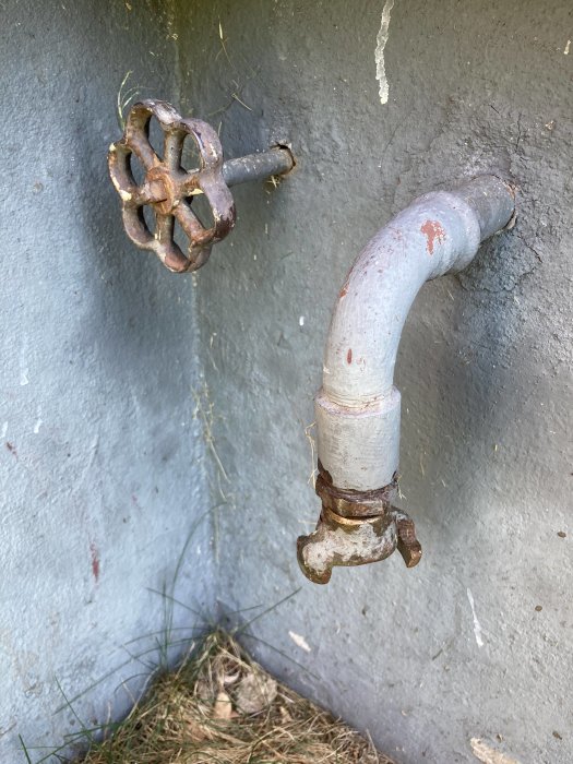 Gammal trädgårdskran med sliten räfflad vred och rostiga beslag monterad på grå vägg.