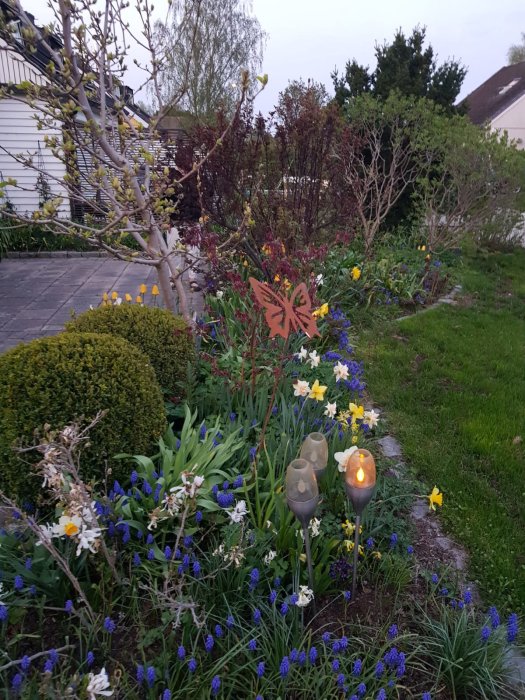 Trädgård i skymningen med blommande hyacinter, påskliljor och andra växter samt dekorativa ljuskällor.