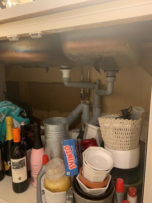 Under-sink cabinet with clutter and visible pipes, potential area for dishwasher installation.