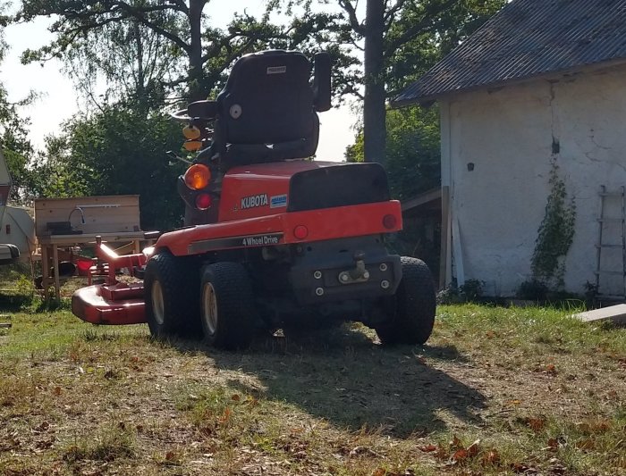 Röd Kubota fyrhjulsdriven gräsklippare parkerad på en stenig och torr gräsmatta med byggnader i bakgrunden.