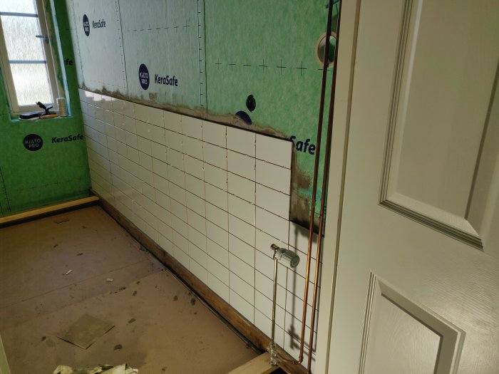 Vägg delvis täckt av nya vita kakelplattor, vattenpass och kakelkryss synliga, orenoverad del av rummet.