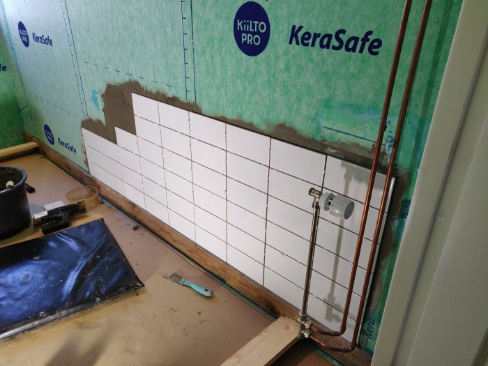 Påbörjad kakelsättning på en vägg med vit kakel halvvägs upp, bredvid rörgenomföringar och en fastkilad lodrät referensplanka.