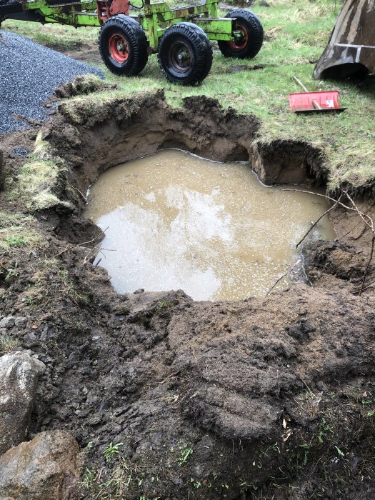 Grävmaskin nära ett öppet provhål fyllt med vatten på kuperad mark, markerar potentiellt grundvatten på en kulle.