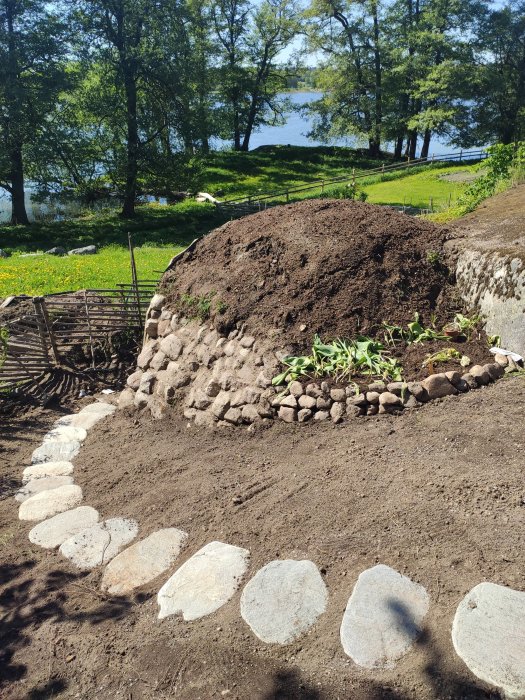 Nyplanterad sluttande trädgård med jordhög, stenar och utsikt över sjö väntande på gräsmatta.