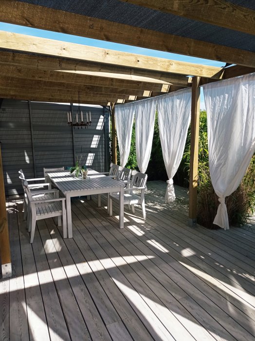 Uteplats med trägolv och alger, vita trädgårdsmöbler under pergola med vita draperier.