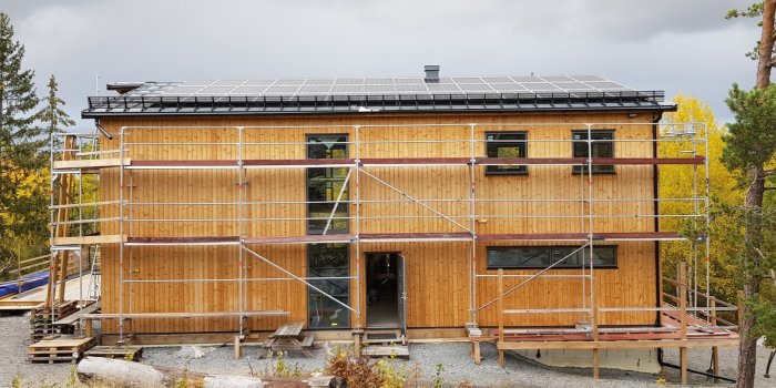 Byggställning i aluminium och stål framför en träbeklädd byggnad med solpaneler på taket, omgiven av träd i höstfärger.
