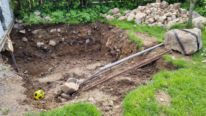 Grävd grop i trädgård för byggprojekt med jordhögar och stenar, metallrör och en fotboll.