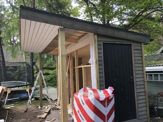 Trädgårdsbod under renovering med nylagt tak och nya klädselbrädor på väggen, omgiven av byggmaterial.